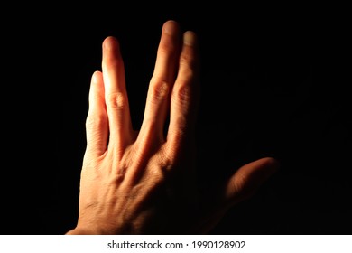 Closeup of left hand back side raised making sci fi salutation gesture isolated on black background. Thumb extended while the fingers are parted between the middle and ring finger. Selective focus.