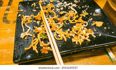 A close-up of leftover noodles on a black plate with scattered toppings and a pair of chopsticks. The composition highlights the casual dining experience, perfect for themes related to food photograph - Powered by Shutterstock