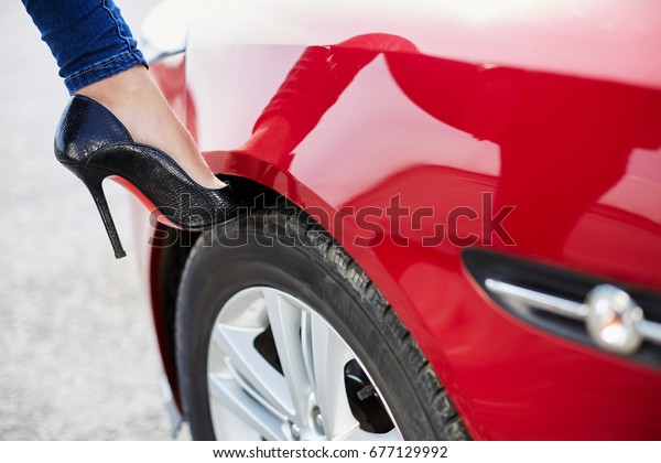 Closeup of leg in jeans and car, driver concept,\
parts of car and body. Leg of woman driver on high-heeled black\
shoes put on wheel of red\
car