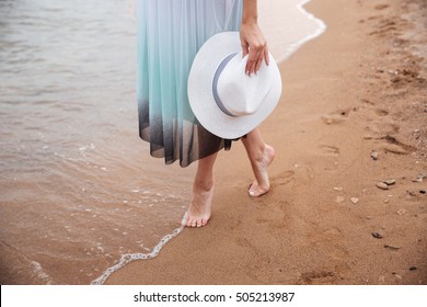 Closeup of legs of young woman in dress holding hat and walking barefoot on the beach