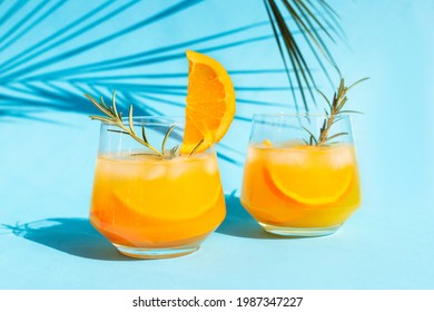 Close-up lemonade, orange cocktail, refreshing summer drink with ice in a glass with a sprig of rosemary on a blue background under a palm leaf on a bright sunny day.