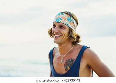 Close-up lifestyle fashion portrait. handsome young man with long curly hair in a bandana smiling.hipster tattoo man.Surfer with a tattoo posing against the backdrop of the ocean. - Powered by Shutterstock