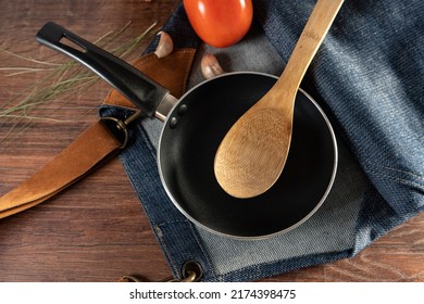 Closeup Of A Light Blue Frying Pan, With A Wooden Spoon And A Jean Apron Resting On A Wooden Table. You Can Also See A Tomato, Garlic And Spices.