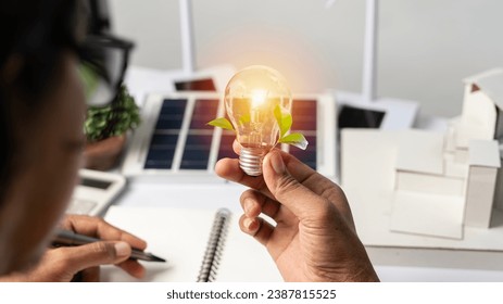 Close-up at a light bulb, An engineer sits holding a light bulb and model of wind turbine. with solar panels and model house on the table. To design use of renewable energy with wind and solar energy.