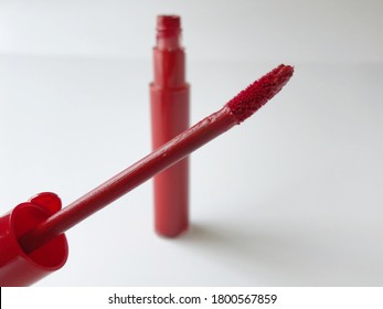 Closeup Of Lip Gloss Wand Applicator Covered In Red Lipstick. The Tube Is In The Background. Isolated On A White Background.