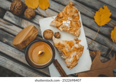 Close-up of a lit candle in an amber glass jar, two slices of apple pie, and a cinnamon stick on a rustic wooden table. Autumn leaves and walnuts enhance the cozy atmosphere near a warm campfire. - Powered by Shutterstock