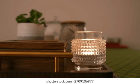 A close-up of a lit candle inside a textured glass holder, creating a warm and cozy ambiance. A small, out-of-focus potted plant is visible in the background.