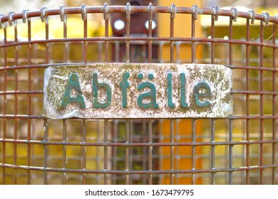 Close-up Of Litter Bin Wire Basket With A Plaque, German Text Sign Abfälle - Waste. Environmental Themed Background With Trash Can, Recycling Bin. Protecting The Environment, Throwing Away Trash Waste