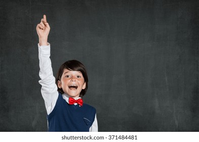 Closeup Of Little Boy With Raised Hand Isolated On Blackboard. Schoolboy Pointing High His Index Finger. Cheerful Cute Boy With Raised Hand Standing Against Black Background.