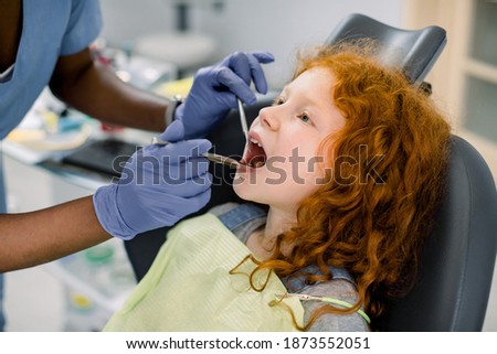 Close-up of little cute curly red haired girl with open mouth during oral checkup at the dentist. Hands of female African American dentist in latex gloves, making teeth examination with tools
