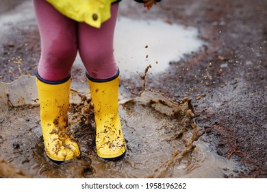 Close-up Of Little Toddler Girl Wearing Yellow Rain Boots And Walking During Sleet On Rainy Cloudy Day. Cute Child In Colorful Clothes Jumping Into Puddle, Splashing With Water, Outdoor Activity