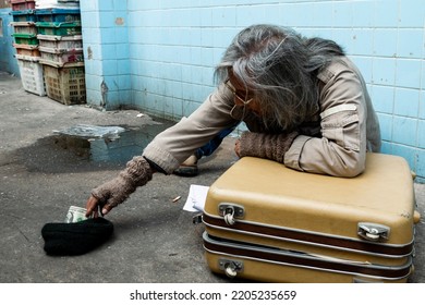 Close-up Of A Long-haired Asian Guy Sitting On A Wall In An Alley, Collecting Money From Passersby For Donations. By Waiting For Help Homeless People Have No Homes To Sleep On The Side Of The Road.