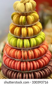 Closeup of lots of multicolored tasty french macarons on tier circle cake tower display stand pyramid-shaped made of clear plastic on many visible levels over blur background, vertical picture 