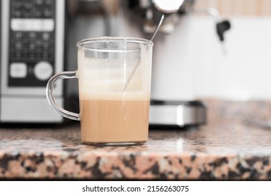 Closeup, Low Angle Of A Glass Coffee Cup With A Teaspoon Inside On A Granite Countertop In A Kitchen, The Coffee Is Half Full. In The Background Out Of Focus Are The Coffee Machine And A Microwave.