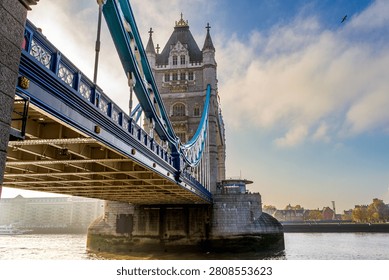 A close-up low angle view highlights the large structural arches and blue suspension cables of Tower Bridge in London. The stone tower rises above the River Thames, while bright morning sun creates golden reflections on the rippling water.