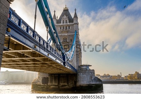A close-up low angle view highlights the large structural arches and blue suspension cables of Tower Bridge in London. The stone tower rises above the River Thames, while bright morning sun creates golden reflections on the rippling water.