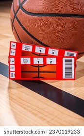 A close-up low angle view of a pair of ticket stubs to courtside seats next to an orange basketball as it sitting a black sideline on wooden floor.