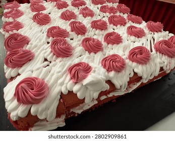 A close-up, low-angle perspective of a large rectangular red velvet or strawberry sheet sponge cake prepared for a catering banquet buffet line. The cake displays clean sponge layers filled with white cream, topped with fluffy white frosting waves, and neatly decorated with structured rows of piped pink cream swirls. Represents commercial bakery art, wedding dessert catering, birthday parties, and festive celebration food.