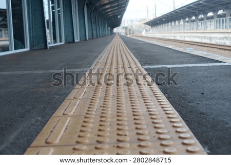 Close-up, low-angle perspective of yellow tactile paving blocks (guiding blocks) for the visually impaired on an empty railway station platform in Indonesia. The composition captures the textured surface of the safety tiles stretching into a vanishing point along the asphalt platform, flanked by modern station infrastructure, glass windows, arched canopy roofs, and empty rail tracks on the side.