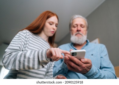 Close-up low-angle view of bearded senior grandfather learning to using mobile phone under guidance of pretty young granddaughter sitting on sofa at home, selective focus.
