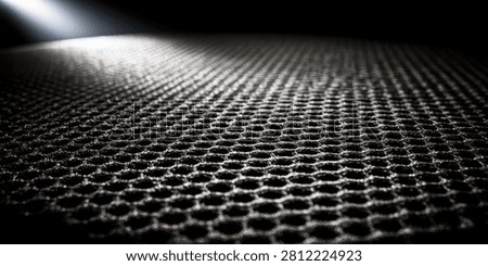 A close-up, low-angle view of a dark, textured surface with a repeating hexagonal pattern, dramatically lit by a bright spotlight from the upper left.