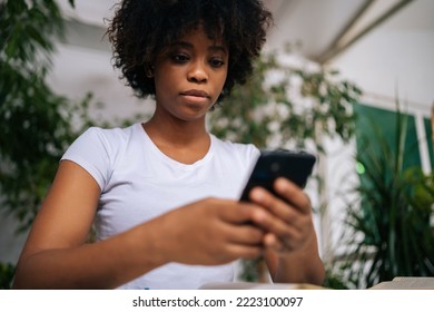 Close-up low-angle view of serious African young woman holding mobile phone in hands, enjoying web surfing, communicating in social network in room with modern biophilia design, selective focus. - Powered by Shutterstock