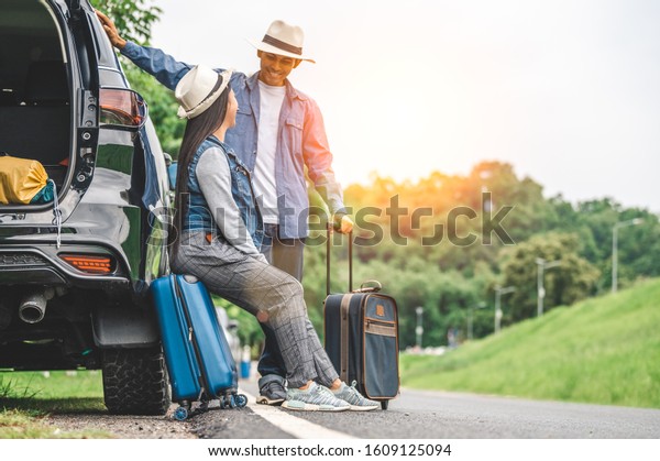 Closeup lower body of Asian couple relaxing on SUV\
car trunk with yellow trolly luggage along road trip with mountain\
hill background. Freedom road life. People lifestyle and\
transportation travel