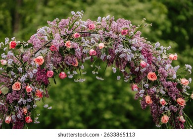 Close-up of a lush wedding floral arch made of lilacs, roses, and purple blossoms, with green foliage and soft natural background.