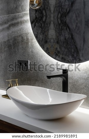 closeup of luxury interior with ceramic washbowl on white countertop, black water tap, grey tile and round mirror on wall in hotel bathroom indoors