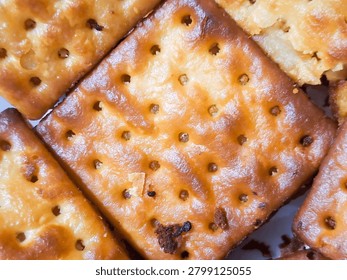 a close-up, macro top-down view of several golden-brown, crispy square crackers arranged tightly together, showcasing their glazed surface texture and evenly spaced docking holes, with a glossy, baked texture. Ideal for snack product packaging, bakery advertising, light breakfast blogging, and savory snack food design concepts.