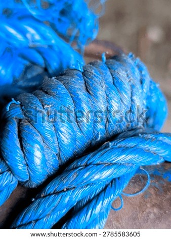  a close-up macro view of thick, vibrant blue synthetic ropes tied into a large, secure knot. The main focus is on the tight coils and twists of the braided strands, which show signs of heavy use, including frayed fibers, dust particles, and slight weathering on the surface. Another layer of the blue rope passes horizontally underneath the main knot, resting against a dark, weathered structure. The background is softly blurred, keeping the emphasis on the detailed texture and vivid color of the cord