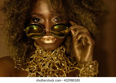 Close-up of magic golden african american female model in massive sunglasses with bright glitter makeup, glossy hairstyle and big red lips posing on the studio background