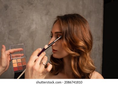 Close-up Of A Makeup Artist Doing Eye Makeup For A Girl Model On A Gray Background In The Studio. Professional Makeup. Place For Text