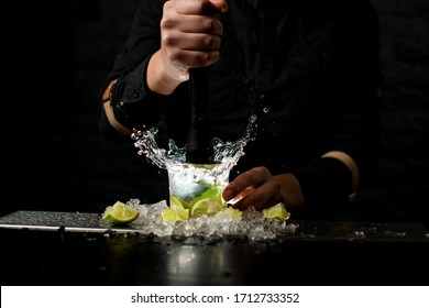 Close-up. Male barman holds black madler in his hand and energetically squeeze slices of citrus to cocktail. Pieces of ice and lime at the bar. - Powered by Shutterstock