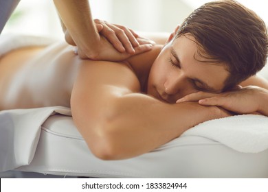 Closeup of male client getting professional body massage in spa salon or wellness center. Young man lying on bed with eyes closed enjoying remedial back massage done by qualified masseur