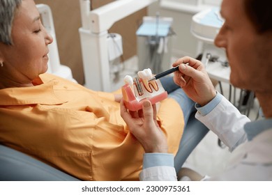 Closeup of male dentist holding tooth model while explaining dental implant surgery to patient in clinic, copy space - Powered by Shutterstock