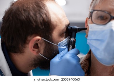 Closeup of male ent specialist with otoscope examining ear of senior woman, both wearing face mask in clinic. Bearded physician using otology tool to check for infections on female pensioner patient. - Powered by Shutterstock