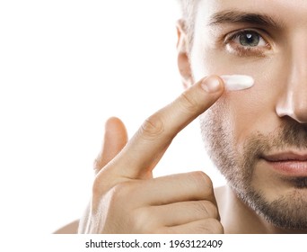 Closeup Of Male Face. Men's Beauty. Young Man Is Applying Moisturizing And Anti Aging Cream On His Face Against White Background