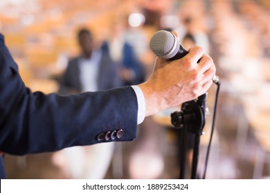 Closeup Of Male Hand In Formal Suit Holding Microphone On Blurred Background Of Conference Hall. Concept Of Speaking During Business Event