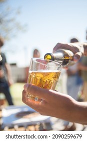 Close-up Of Male Hand Holding Mug And Pouring Cold Beer In It. Fresh Alcohol Beverage In Glass, Friends Of Different Nationalities And Pizza In Background. Party, Friendship Concept