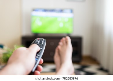 Closeup Of A Male Hand Holding Remote Control With His Feet Crossed In Front Of A Large Television Set Sitting In His Living Room And Watching Football. Changing The Channel