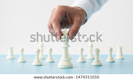 Closeup of male hand holding white chess figure of king positioned in front of the other figures on a blue desk in a conceptual image.