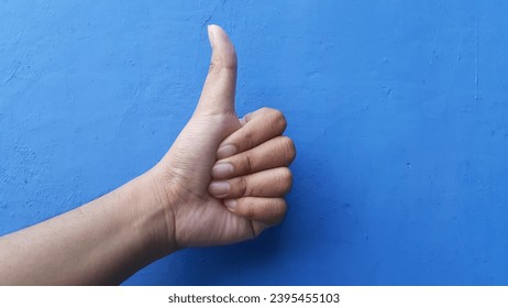 Closeup of male hand showing thumbs up sign against blue wall background. - Shutterstock ID 2395455103