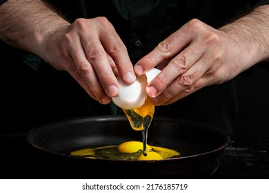 Closeup Of Male Hands Cracking Egg For Scrambled Eggs Omelette. Baker Man Wearing A Black Outfit In Darkness. Eggshell, Egg White, Yolk.