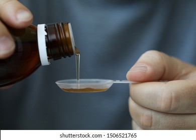 Close-up Of Male Hands Holding Holding Bottle Of Cough Expectorant Syrup And Pouring It With Measuring Spoon. Ill Man Taking Medicine. Healthcare And Treatment Concept. Blurred Background