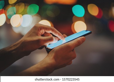 Closeup of male hands using modern smartphone device outdoors, bokeh light on the background, Man typing an sms message on her cellphone while walking at night city streets - Shutterstock ID 1570223545