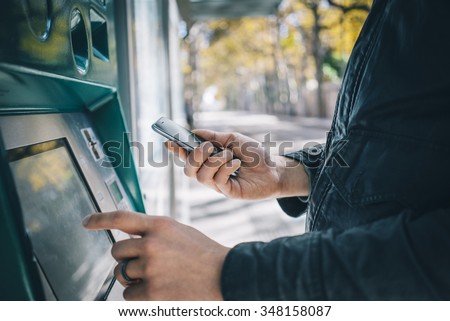 Closeup of male hands using smart phone while typing on ATM, bank machine