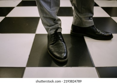 Close-up Of Male Legs In Black Leather Shoes On A Black And White Tile. Groom In Gray Trousers And Black Leather Shoes, Legs Close-up