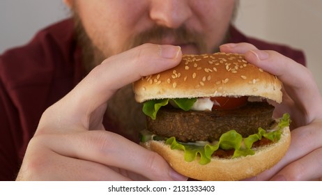 Close-up Of Male Mouth Eating Vegan Burger. Man Eating A Hamburger. Fast Food Eats. Burger In Male Hands. A Bearded Man Eats A Delicious Hamburger Or Cheeseburger.