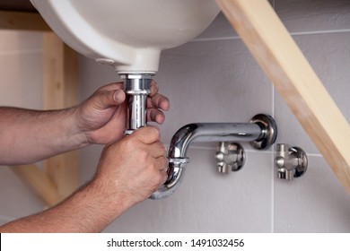 Closeup Male Plumber Worker In Blue Denim Uniform, Overalls, Fixing Sink In Bathroom With Tile Wall. Professional Plumbing Repair Service, Installation Of Water Pipes, Man Mounted Sewer Drain